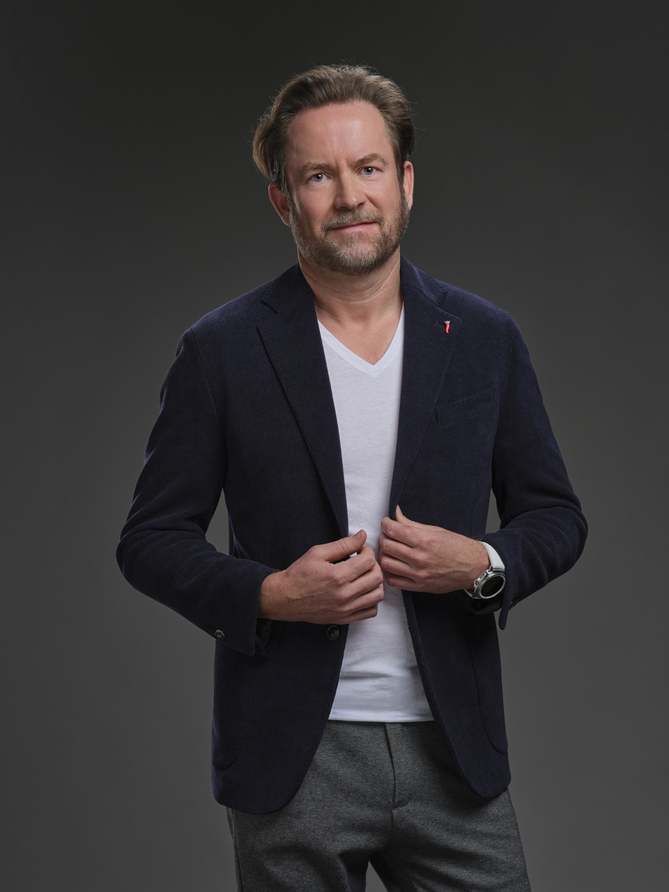 Markus Miller im dunkelblauen Sakko und weißem V-Shirt vor grauem Studiohintergrund